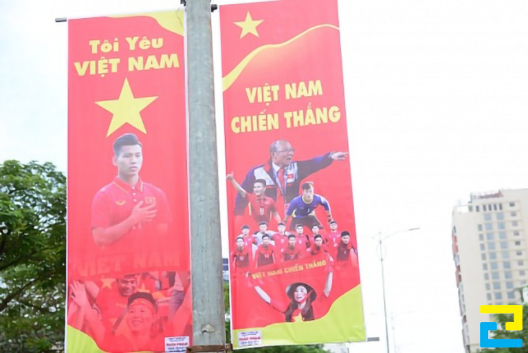 Mẫu 7: Băng rôn bóng đá khổ dọc được thiết kế với kiểu chữ màu vàng, nền đỏ, có hình ảnh của người cổ vũ - đội tuyển - huấn luyện viên